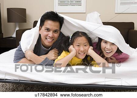 Picture - Mother, father and daughter lying down on bed covered with bed sheet. Fotosearch