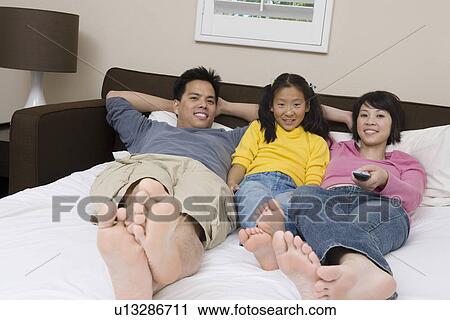Stock Image - Mother, father and daughter lying down on bed and watching TV. Fotosearch