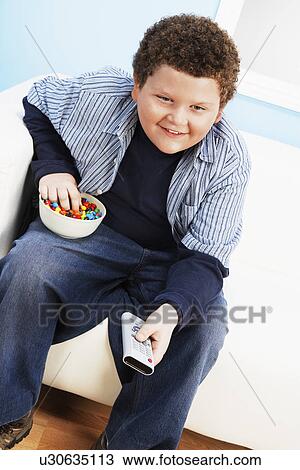 Stock Image - Overweight boy (13-15) Eating Junk Food, holding remote control. Fotosearch