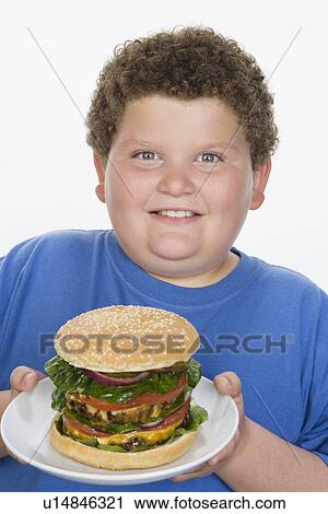 Stock Image - Overweight boy holding plate with big cheeseburger. Fotosearch