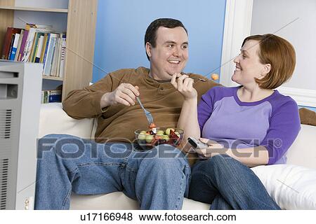Stock Photo - Overweight couple eating fruit on sofa. Fotosearch