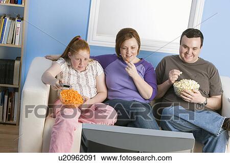 Stock Image - Overweight family watching television on sofa. Fotosearch