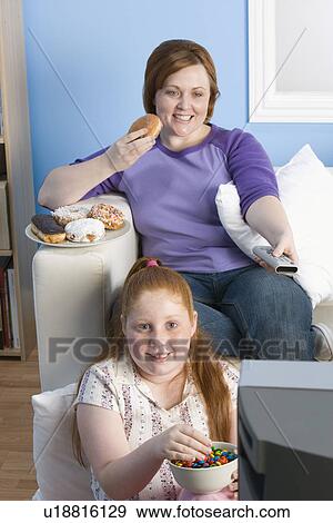 Stock Photo - Overweight girl and mother watching television, eating. Fotosearch