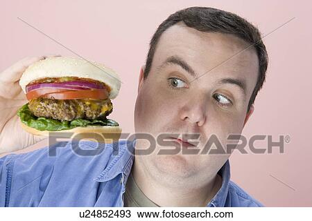 Stock Image - Overweight mid-adult man holding hamburger. Fotosearch