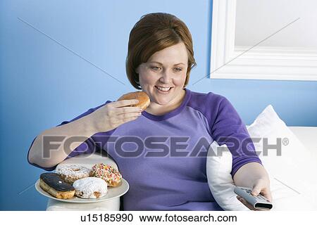 Stock Image - Overweight woman watching television, eating. Fotosearch