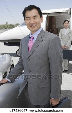 Stock Photo - Portrait of mid-adult businessman standing in front of convertible, mid-adult businesswoman in background.. Fotosearch