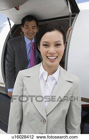 Stock Image - Portrait of mid-adult businesswoman standing in front private jet, colleague behind.. Fotosearch