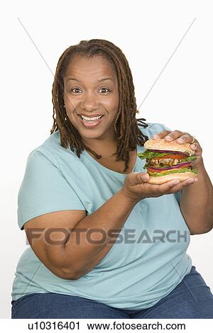 Stock Image - Portrait of Mid-adult overweight woman holding big cheeseburger and smiling. Fotosearch