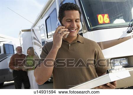 Picture - Salesperson with clipboard using mobile phone, customers in background, portrait. Fotosearch