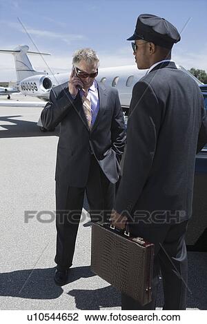 Stock Image - Senior businessman standing outside private jet and talking on phone, Chauffeur holding his suitcase.. Fotosearch