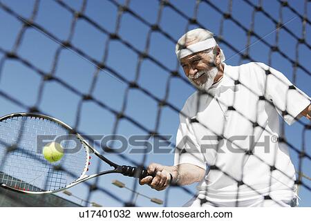 Stock Image - Senior man hitting tennis ball forehand near tennis net. Fotosearch