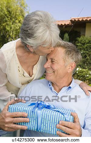 Stock Image - Senior woman giving husband birthday present and kissing him on forehead. Fotosearch