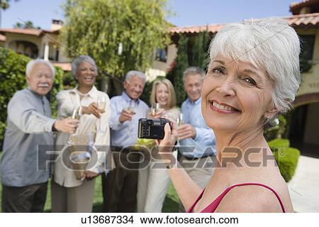 Picture - Senior woman photographing friends in garden while raising toast. Fotosearch