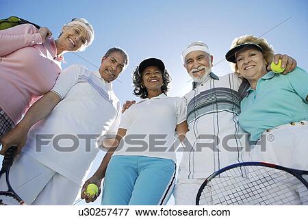 Stock Photo - Seniors standing in half circle, holding rackets and balls, low angle view. Fotosearch
