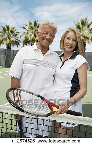 Stock Photograph - Smiling Couple standing on Tennis Court, arms around, holding rackets. Fotosearch