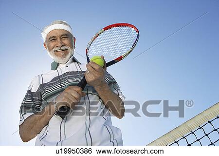 Stock Photograph - Smiling Man holding racket and ball, standing above net, low angle view. Fotosearch