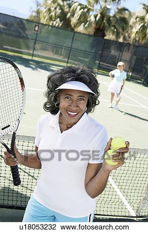 Stock Image - Smiling senior Woman holding tennis balls and racket on tennis court. Fotosearch