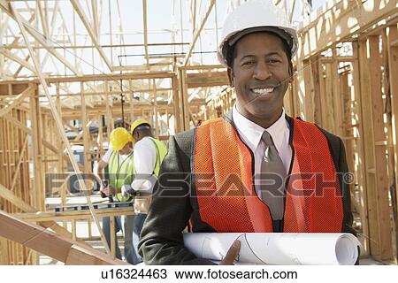 Stock Image - Smiling Surveyor in hard hat with building plans on Construction Site. Fotosearch