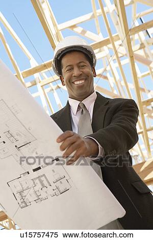 Stock Photography - Smiling Surveyor in hard hat with building plans on Construction Site, low angle view. Fotosearch