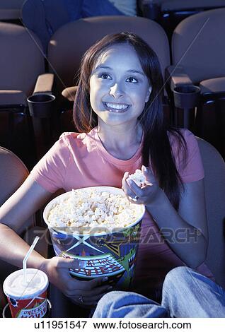 Stock Photo - Smiling young woman Eating Popcorn, watching movie in Theatre. Fotosearch