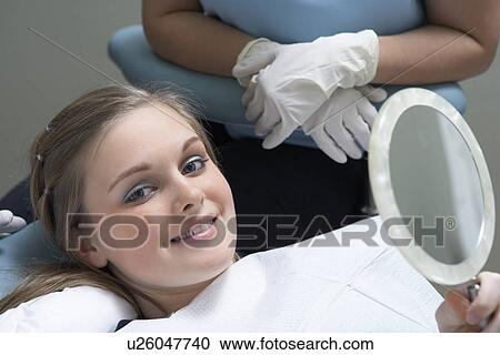 Stock Image - Teenage girl (13-16) holding mirror at dentists, (portrait). Fotosearch
