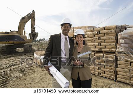 Two architects standing on construction site holding blueprints View Large Photo Image Stock Image - Two architects standing on construction site holding blueprints. Fotosearch