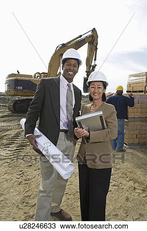 Two architects standing on construction site holding blueprints View Large Photo Image Stock Image - Two architects standing on construction site holding blueprints. Fotosearch