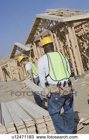 Stock Image - Two construction workers carrying wooden plank on site. Fotosearch