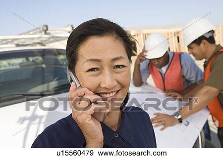 Two construction workers looking at blueprints and architect talking on phone on construction site View Large Photo Image Stock Photo - Two construction workers looking at blueprints and architect talking on phone on construction site. Fotosearch