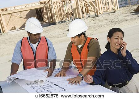 Stock Photo - Two construction workers looking at blueprints and architect talking on phone on construction site. Fotosearch