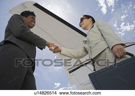 Picture - Two mid-adult businesswomen shaking handsin front of private plane, low angle view.. Fotosearch