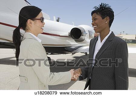 Stock Photo - Two mid-adult businesswomen shaking hands in front of private plane on runway.. Fotosearch