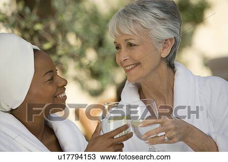 Stock Image - Two women in bathrobes, toasting drinks. Fotosearch
