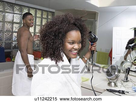 Stock Photography - Woman blow-drying hair with boyfriend standing behind in bathroom. Fotosearch