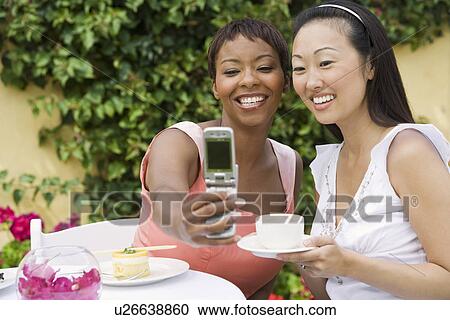 Stock Image - Women photographing themselves at outdoor table. Fotosearch