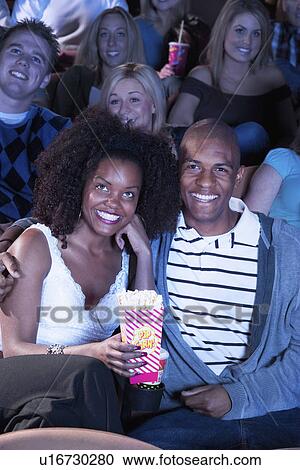 Young Couple sitting in theatre, arms around, Watching Movie, eating popcorn View Large Photo Image Stock Image - Young Couple sitting in theatre, arms around, Watching Movie, eating popcorn. Fotosearch