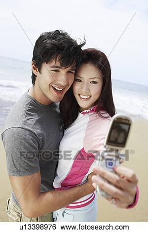 Stock Photograph - Young couple taking picture with cell phone on beach. Fotosearch