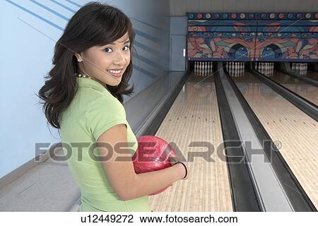 Stock Image - Young woman at bowling alley holding ball, portrait. Fotosearch