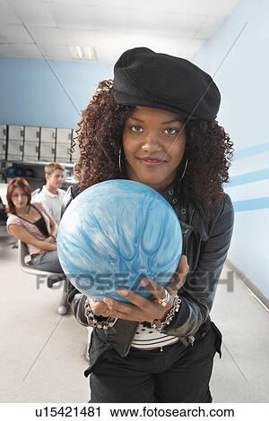Stock Image - Young woman at bowling alley holding ball, portrait. Fotosearch