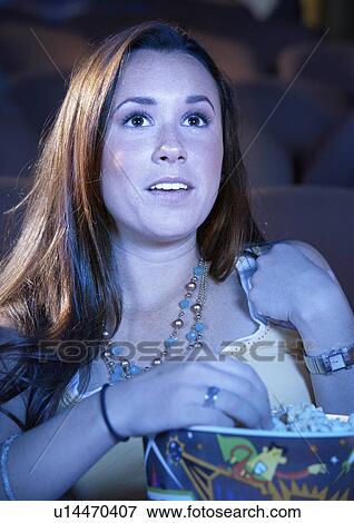 Stock Photo - Young woman Eating Popcorn, watching movie in Theatre. Fotosearch