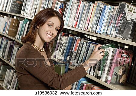 Stock Image - Young woman in Library, selecting book from shelf. Fotosearch