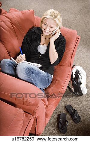 Stock Photo - Young woman on sofa using telephone and writing, elevated view. Fotosearch