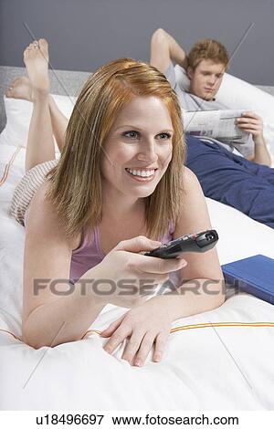 Young woman using remote control lying in bed next to boyfriend View Large Photo Image Stock Photo - Young woman using remote control lying in bed next to boyfriend. Fotosearch