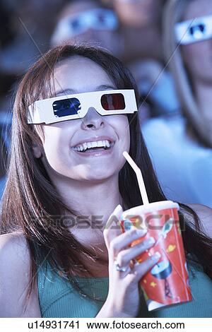 Stock Image - Young woman wearing 3-D glasses watching 3-D movie in theatre, drinking soft drink. Fotosearch