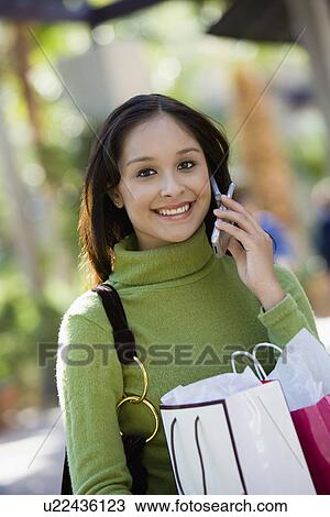 Stock Image - Young woman with shopping bags using mobile phone, portrait. Fotosearch