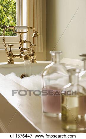 Stock Photo - Bottles of Bath Oils on edge of bathtub filled with bubbles, water running from tap. Fotosearch