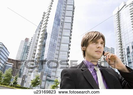 Stock Image - Business man using mobile phone, standing outside office buildings. Fotosearch