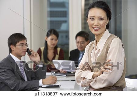 Stock Photo - Business woman sitting at conference table with colleagues in background, portrait. Fotosearch