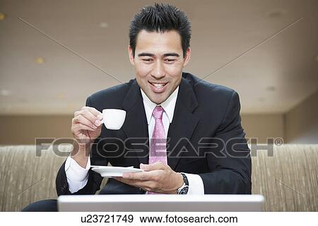 Businessman holding cup of coffee, looking down at laptop in office View Large Photo Image Stock Photo - Businessman holding cup of coffee, looking down at laptop in office. Fotosearch