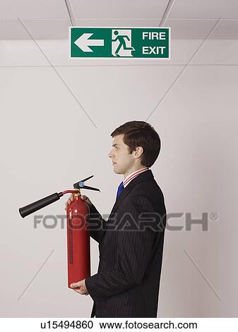 Stock Image - Businessman holding fire extinguisher under exit sign. Fotosearch
